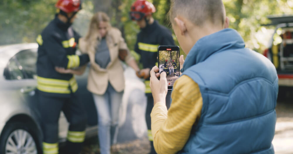 Boy records car accident scene as firefighters assist injured woman — potential witness capturing evidence after a crash.